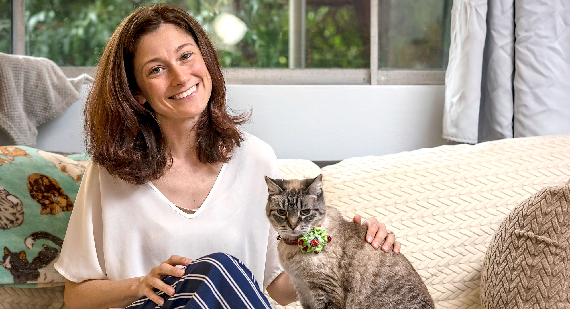 Natasha is wearing a white blouse and blue pants with white stripes. She is sitting on a cream couch with her tabby cat next to her.