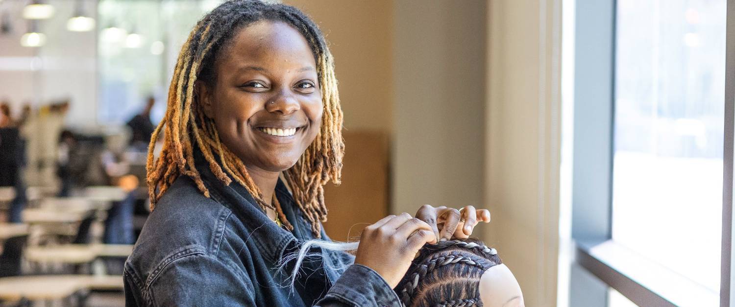 A cosmetology student wearing a jean jacket is practicing braiding hair on a mannequin head.