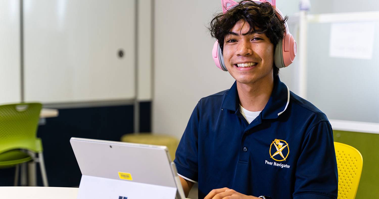 A student in a blue polo shirt wears headphones while working on a laptop