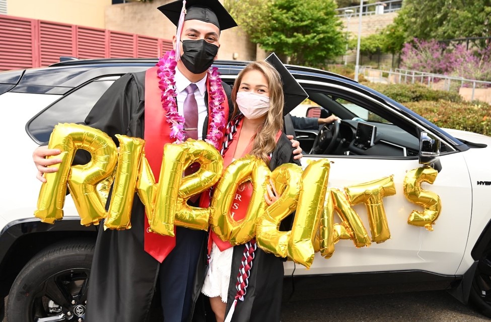 Two nursing students wear graduation caps and gowns at hold gold balloons that say R N and 2021