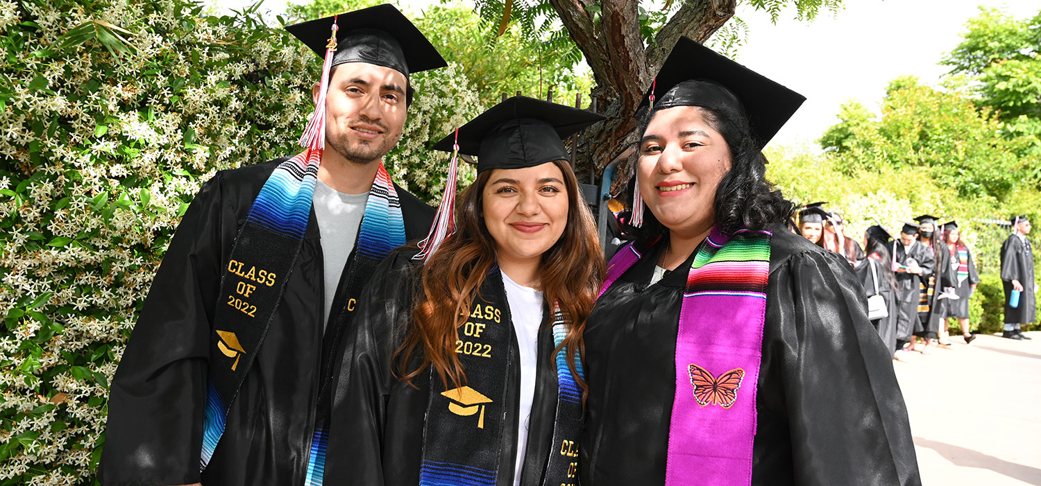 Three students in caps and gowns stand by a tree before the 2022 City College commencement