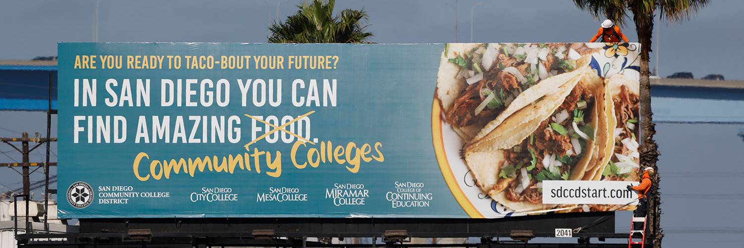 Workers install a billboard. The billboard shows a plate of taco. Text reads Are you ready to taco bout your future In San Diego you can find amazing food which is crossed out and replaced with community colleges. 