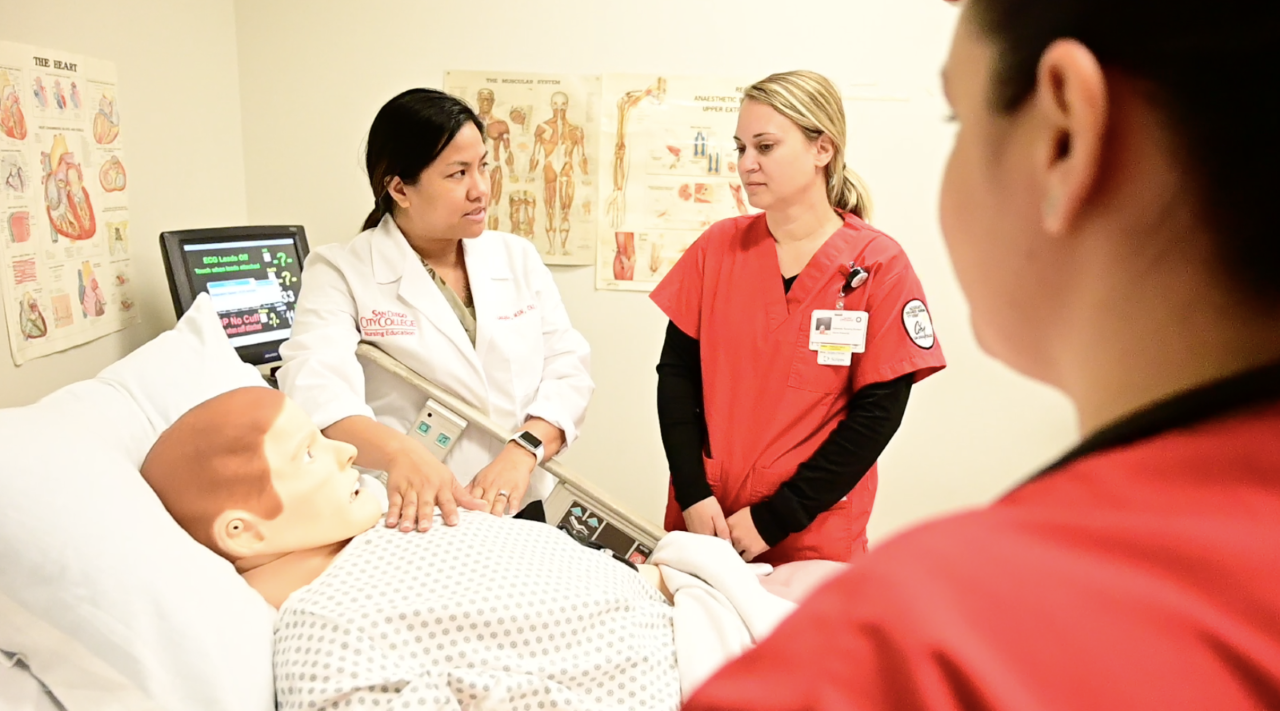 An instructor is wearing a white doctors coat while two students are wearing red hospital scrubs. They are standing over a dummy in a hospital bed.