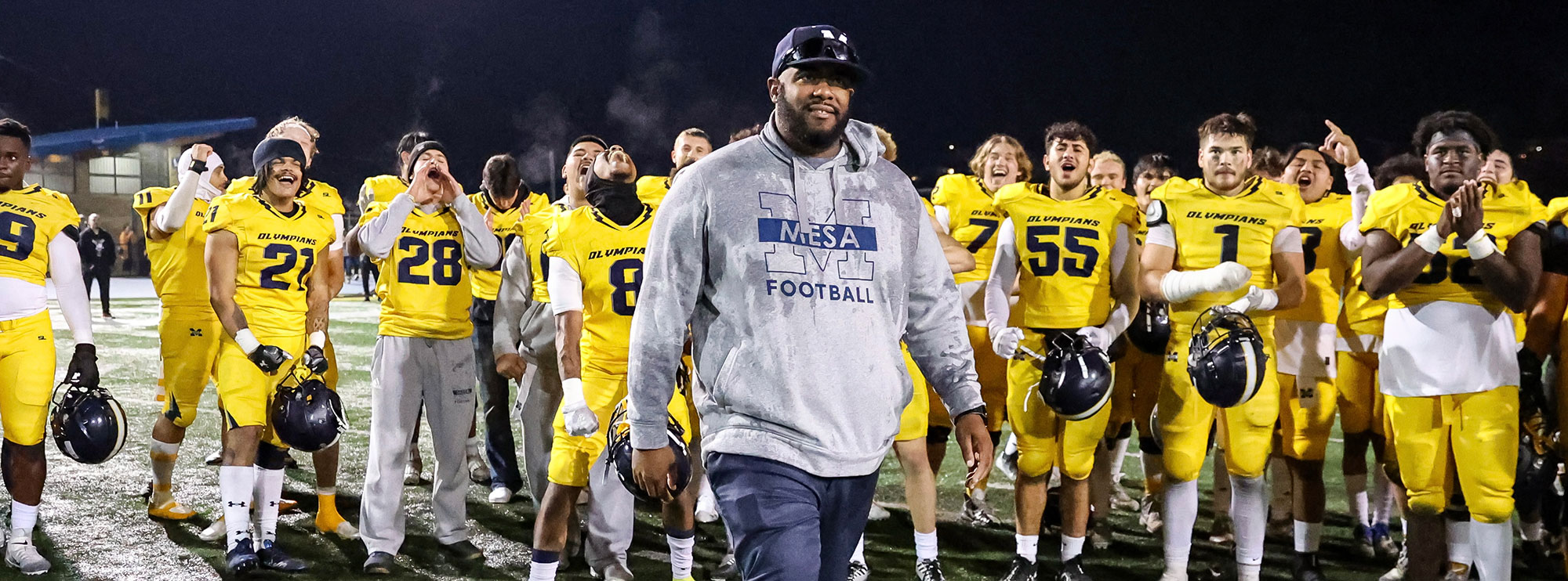 Coach Gary Watkins walks on the football field at night after a game. Football players are in yellow shirts behind him applauding.