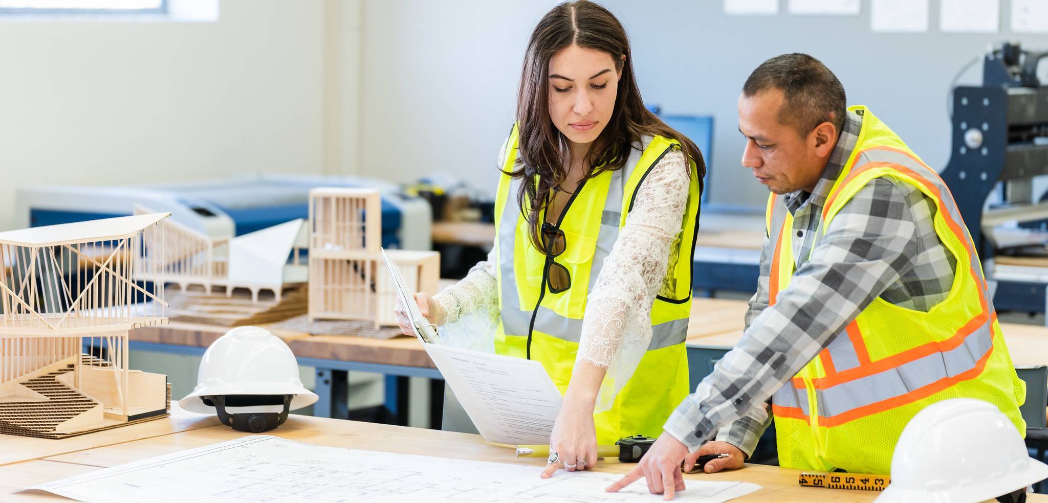 A man and a woman are looking at architecture plans at a work table. They are wearing yellow reflective vests and two white hard hats are on the table. 
