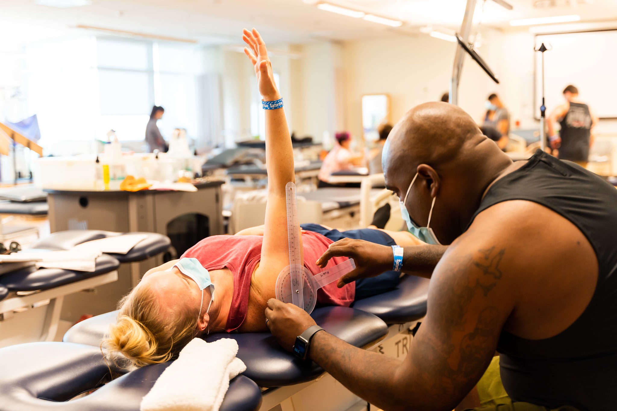 A woman laying on a physical therapy table lifts her arm while a therapist measures her the range of motion in her shoulder