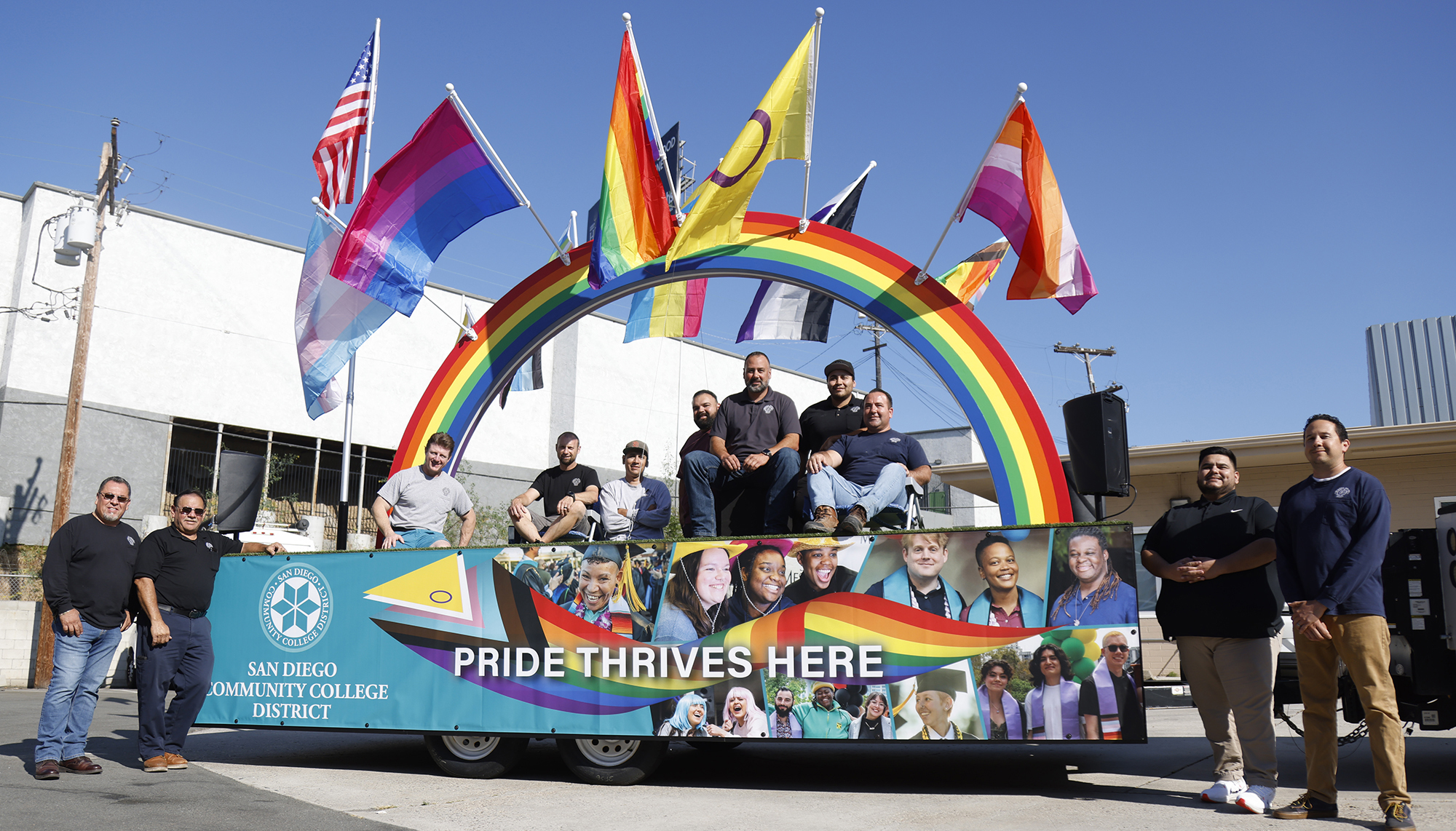 11 people are around the Pride Parade Float. The float has a giant rainbow that extends from the front to the back and has 8 various pride flags that hang on the side of the rainbow. 