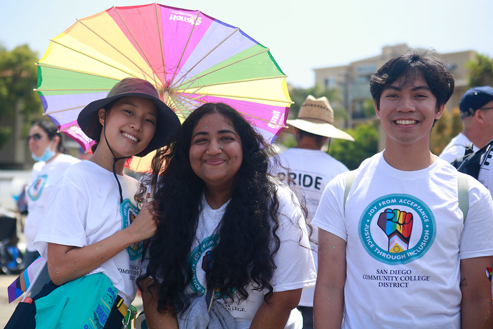 Three friends take a group photo under a rainbow umbrella at the 2022 Pride Parade