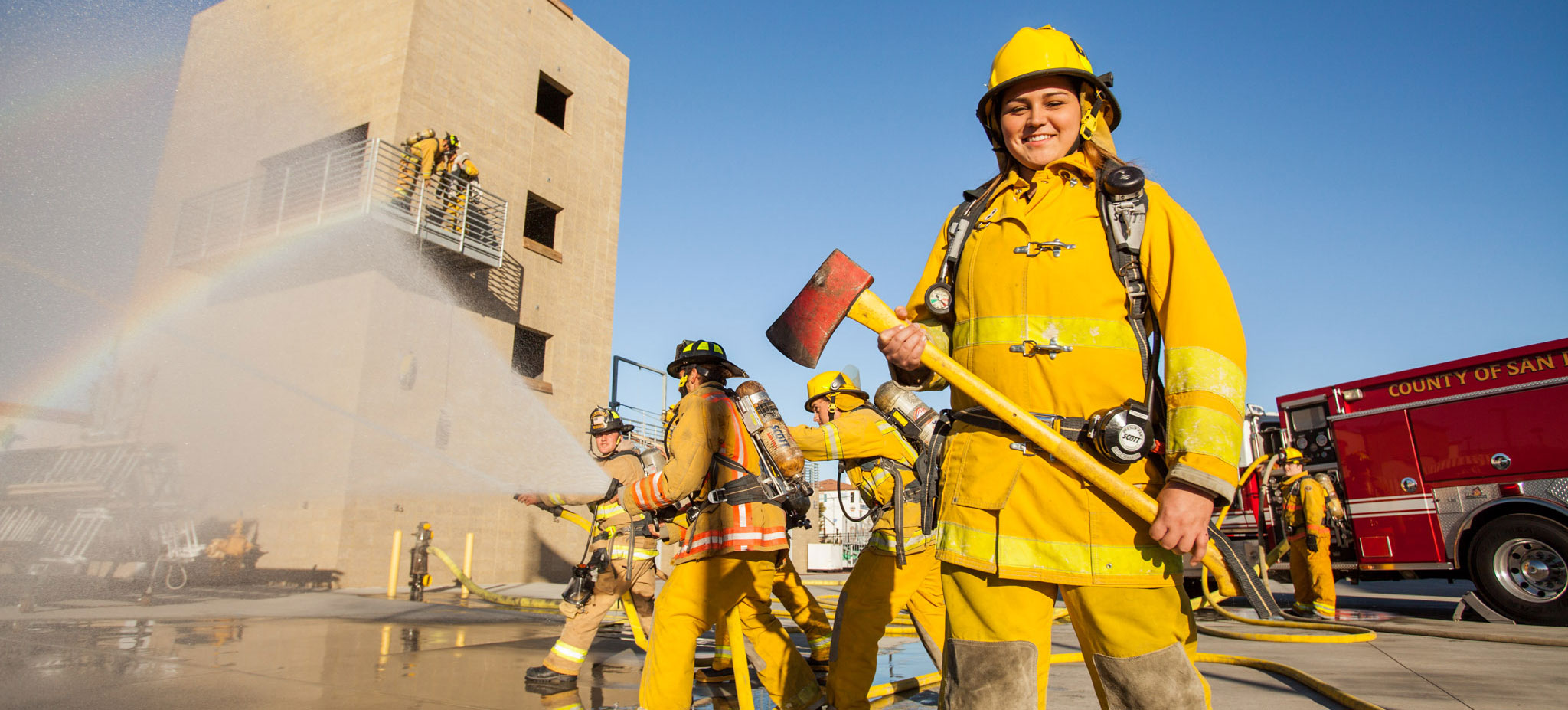 A firefighting student is wearing yellow protective fire gear and holding an axe. There are three fire students behind her in yellow gear using fire hoses