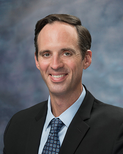 Portrait photo of Gregory Smith wearing a black jacket and a blue shirt and tie