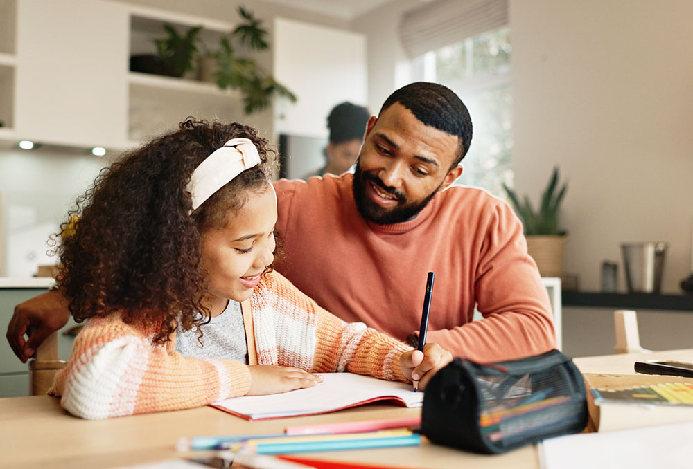 A father looks over his daughter's shoulder as she works on her homework at the kitchen table