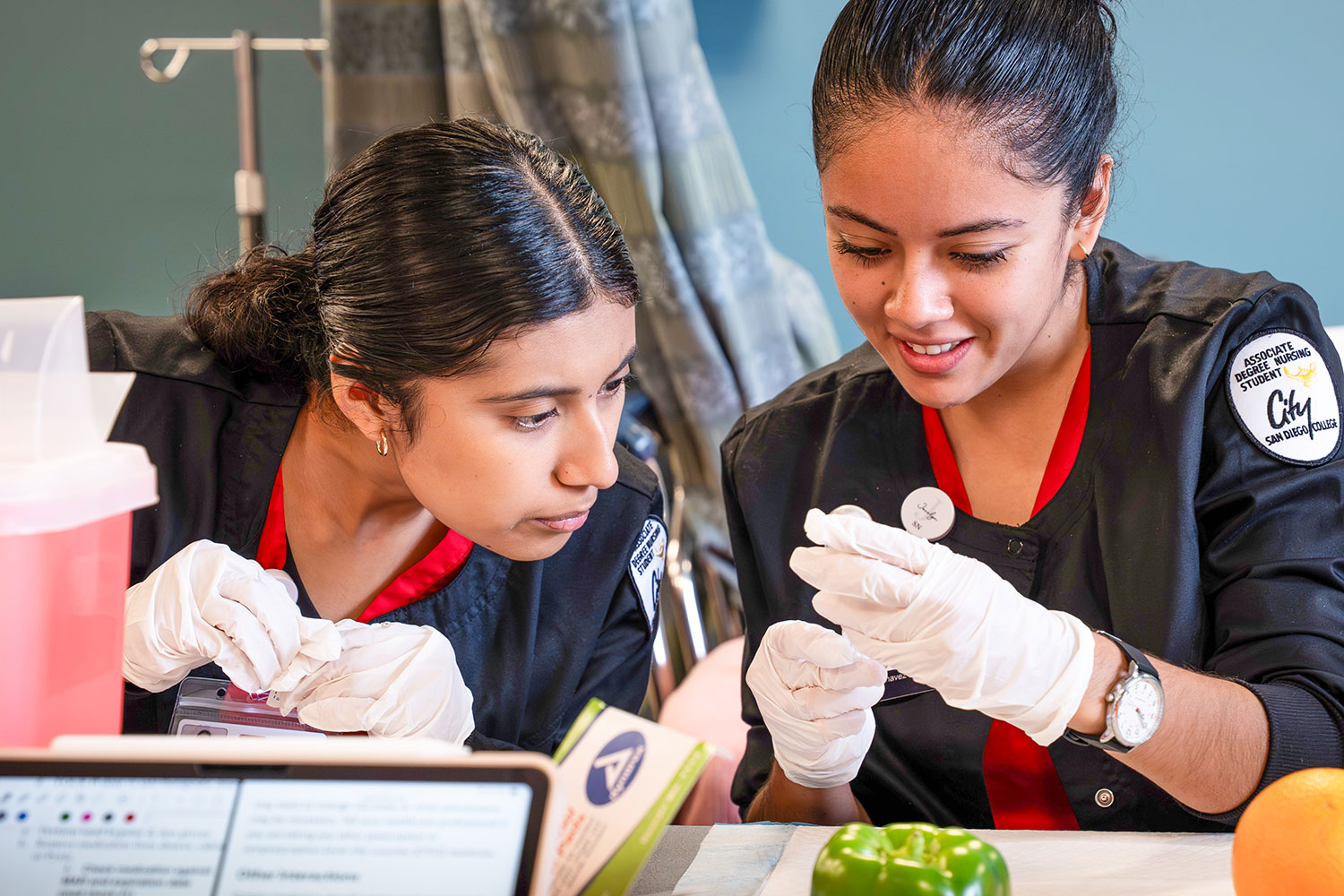 Two City College nursing students in black and red scrubs look closely at a vial.
