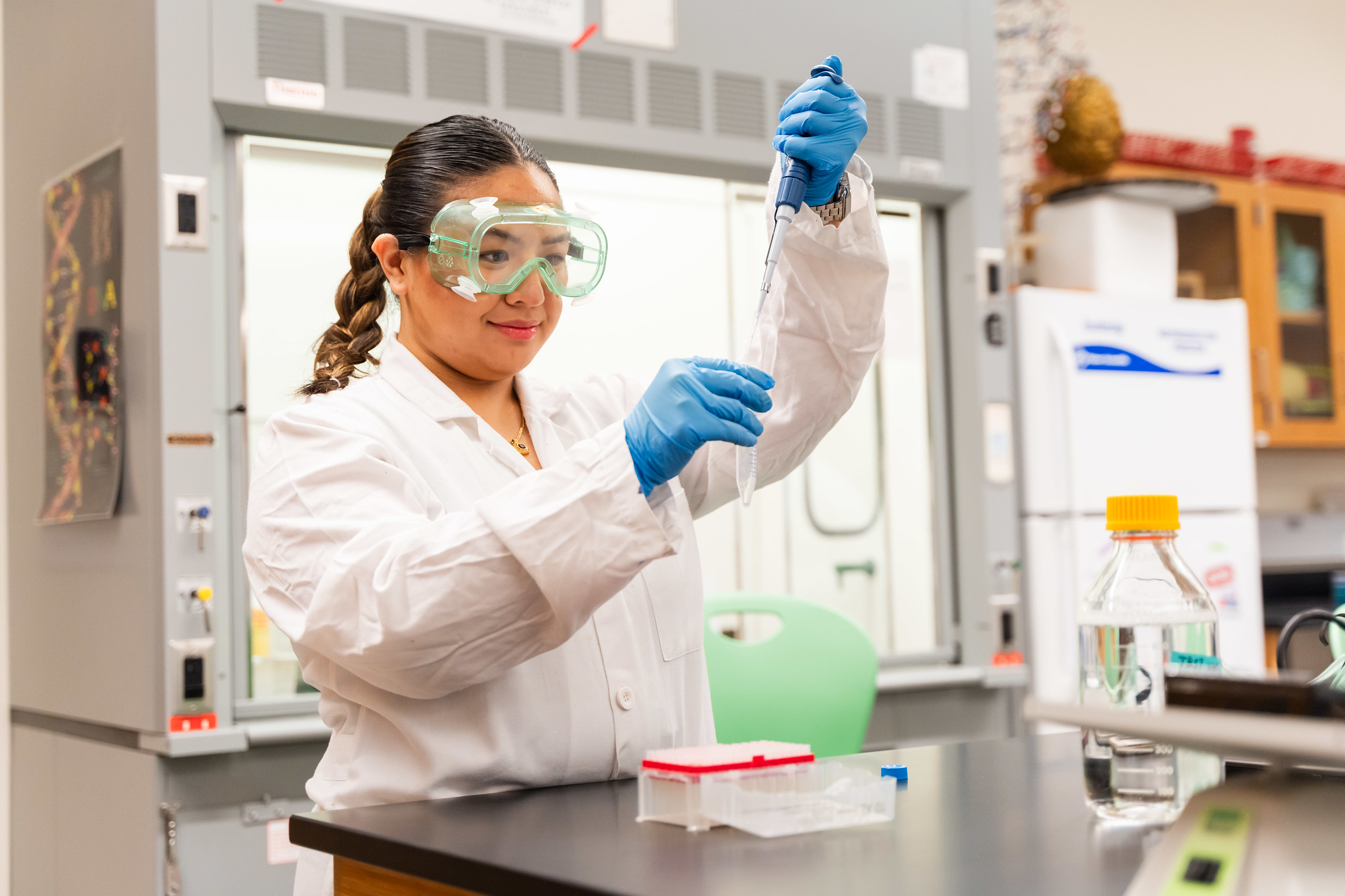 Young woman with dark brown hair pulled back into a braid in a white lab coat, blue gloves, and clear goggles, dropping liquid into a test tub in a lab.