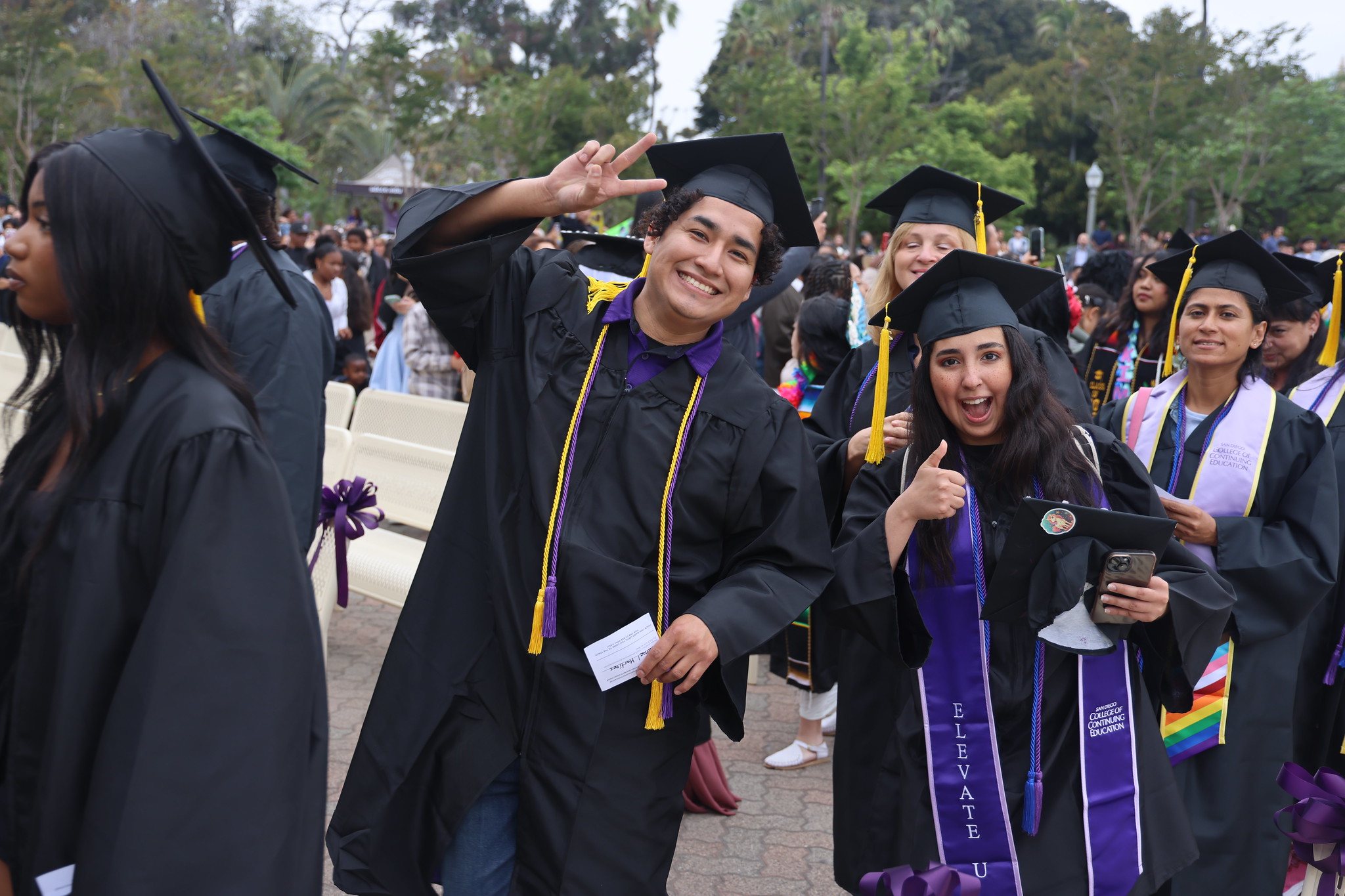 
Two graduates give a peace sign and a thumbs up while waiting in line to receive their certificates.

