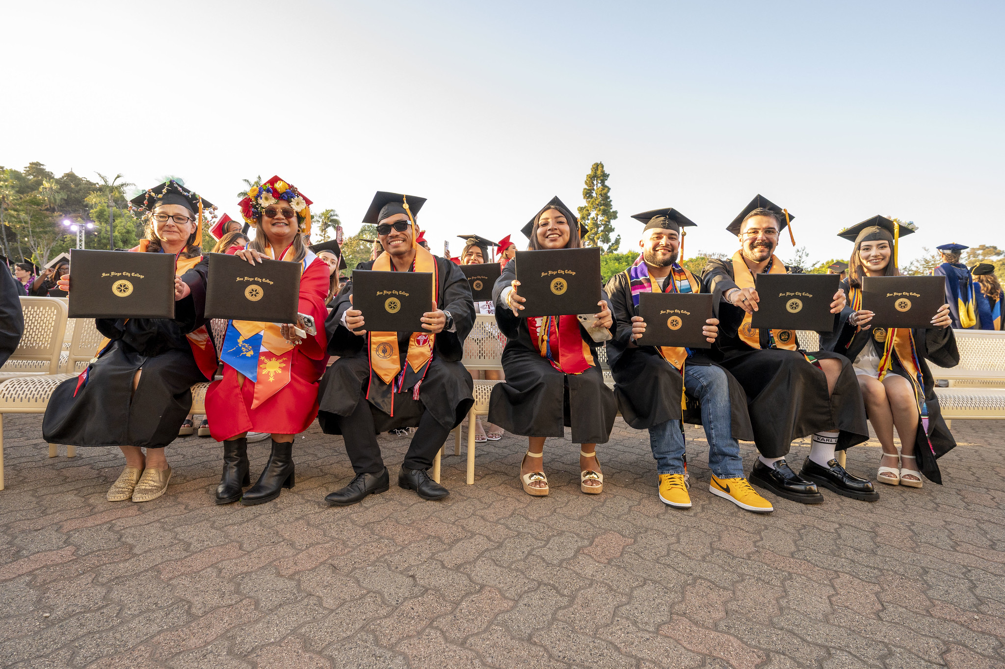 
Seven grads sitting on a bench all holding up their degrees.
