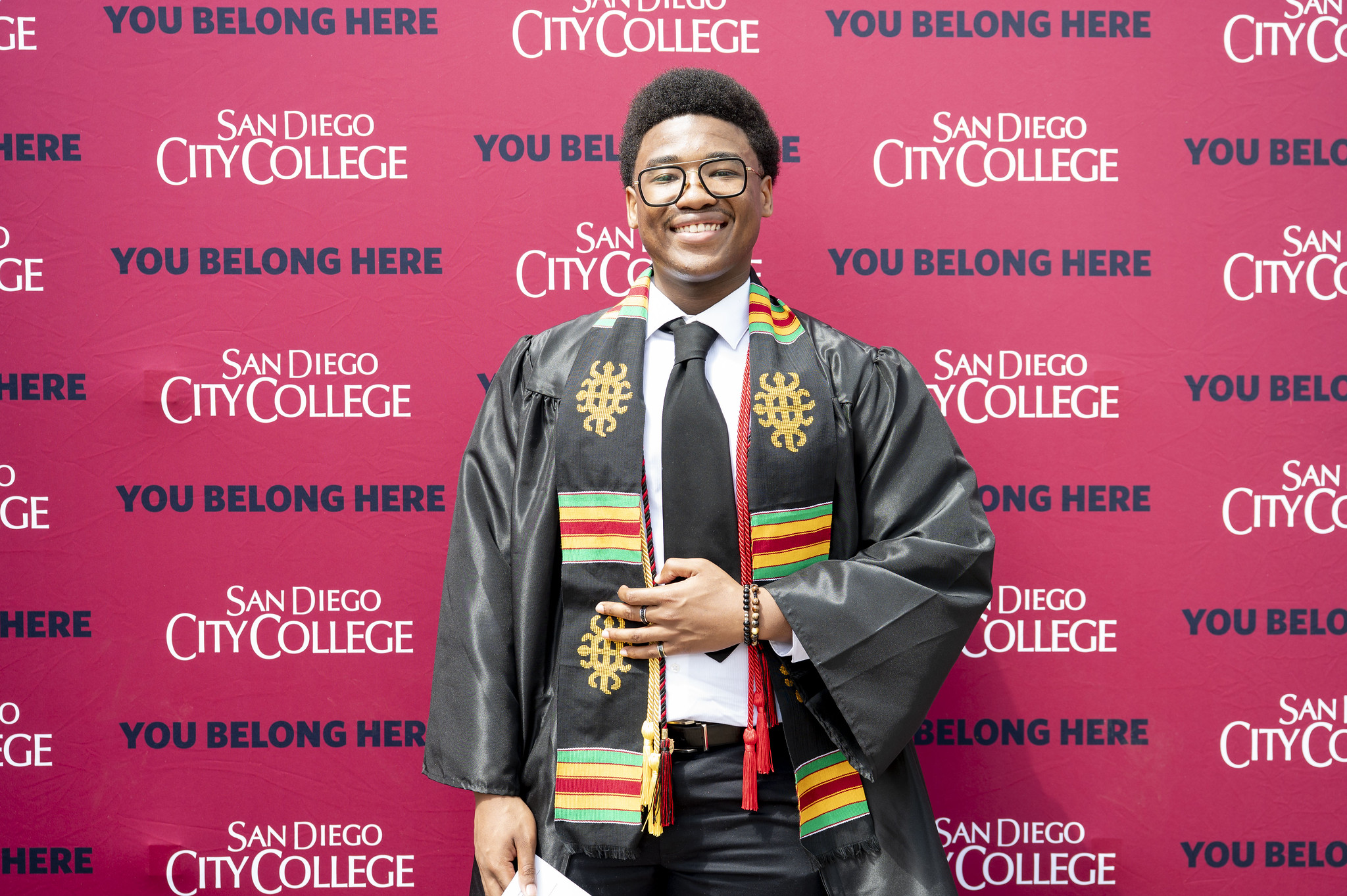 
A student in a graduation gown in front of a red background with the City College logo.
