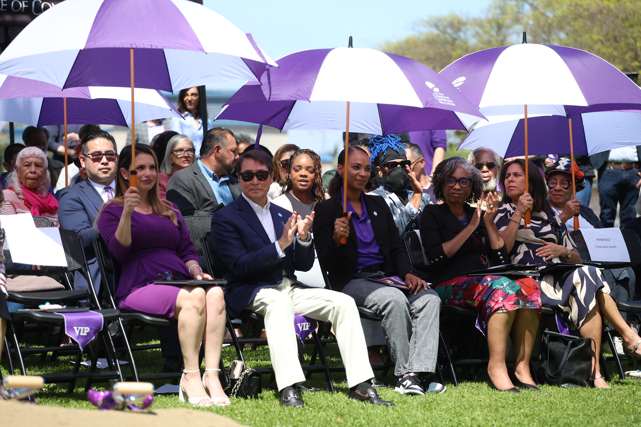 
Attendees hold matching purple and white large golf umbrellas the shade the audience
