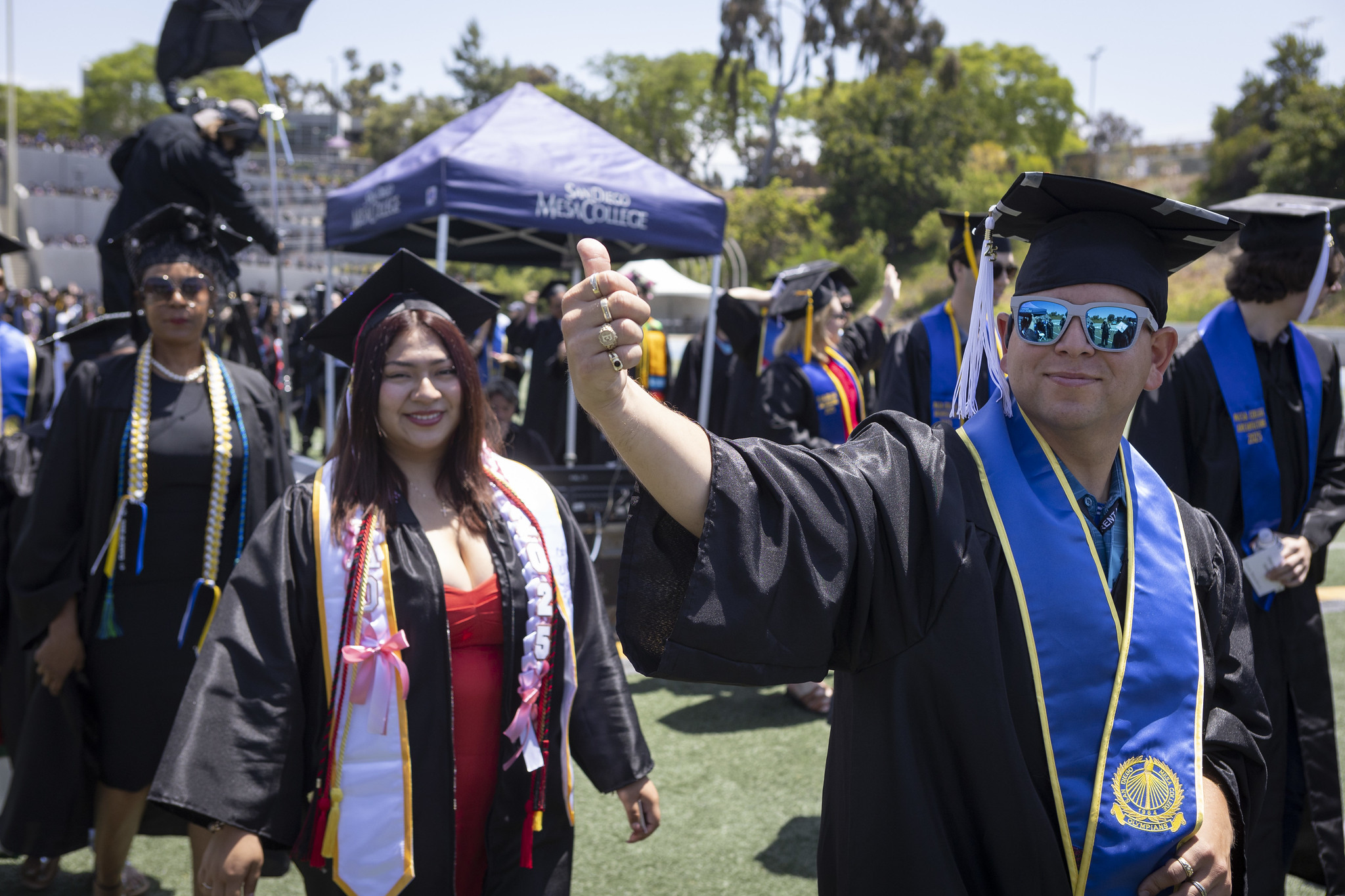 
Three graduates walking to their seats. The man in front gives a thumbs up to the audience.
