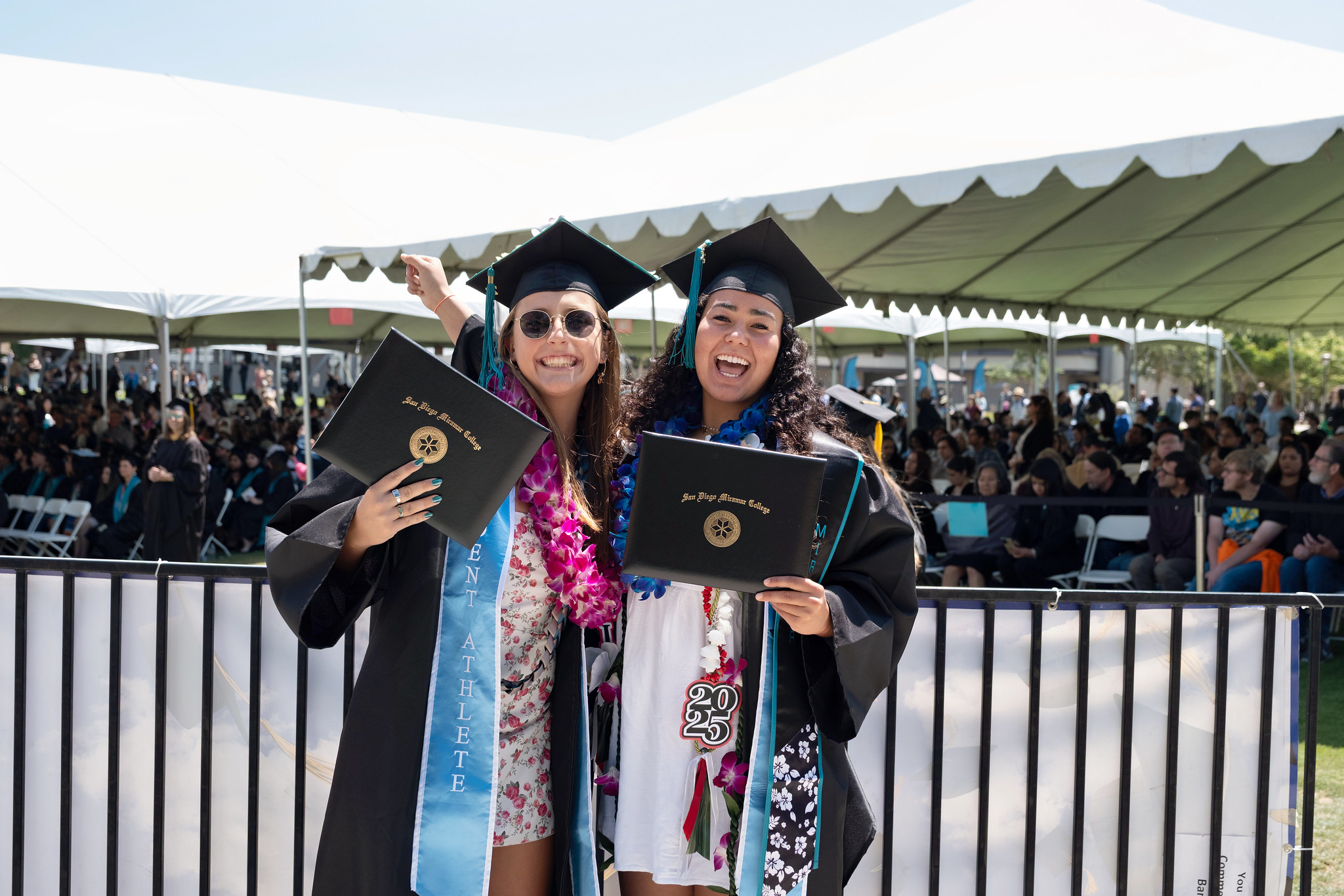 
Two students in black caps and gowns hold up their degrees as they leave the stage at commencement.
